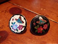 Pair of cloisonné butterfly button brooches with colorful floral designs on wooden surface.