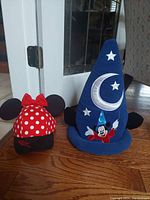 Both Disney children's hats together placed on wooden floor in front of a glass door and floral wallpaper, showing Minnie Mouse and Mickey Mouse designs.