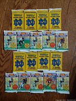 Top down view of 18 unopened trading card packs laid out on a wooden floor. Packs include yellow University of Notre Dame 1st Edition packs and green/white Upper Deck NFL football trading card packs.