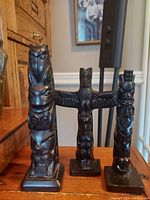 Photo of three black cultured stone North West First Nations replica totem poles arranged on a wooden surface showing front view and details of carved animal faces and tribal symbolism.
