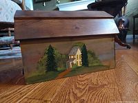 Forward view of wooden mailbox showing hand painted folk art house scene with trees on front panel.
