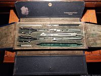 Open drafting set showing multiple metal drafting tools including compasses and dividers arranged in a cloth-lined black case with a worn spot inside the lid.
