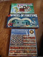Photo showing three boxed items: a Wheel of Fortune 48 Games Kit box on top, a Smithsonian jigsaw puzzle box below, and a partial view of the Wheel of Fortune game box behind them.