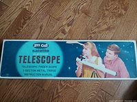 Original SkilCraft Satellite 472 telescope box showing front design with title and children using telescope graphic, on wood floor background.