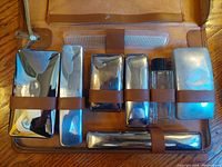 Top view of the kit interior showing multiple polished stainless steel containers secured by leather straps, and a clear bottle with black cap. Includes razor and brushes.