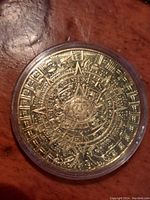 Gold-toned coin with Aztec Sun Stone design shown face up in clear plastic case on wood surface.