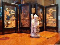 Front view of the four-panel cork art folding screen and small porcelain God of Longevity statue on wooden surface under warm lighting.