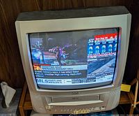 Front view of a 13-inch RCA TV/VCR combo unit powered on, displaying a news broadcast with visible control buttons and tape deck slot.