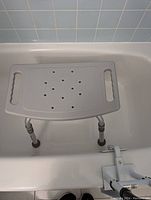 Adjustable shower seat placed inside a bathtub showing its plastic seat with drainage holes and metal adjustable legs with rubber tips for stability.