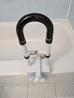 Photo of the adjustable tub handrail clamp attached to a bathtub edge, showing the black arched handle and white clamp mechanism.