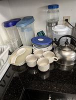Photo showing a silver metal kettle with black handle and lid knob, four white ceramic teacups, one mug, two tall plastic food storage bins with blue and gray lids, a ceramic spoon rest, several glass jars including a large jar with blue lid, and a white dish drainer.