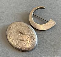 Two vintage sterling silver brooches on gray background, one oval with detailed floral engraving and one crescent shaped with smooth finish.