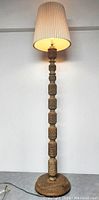 Full view of the carved teak floor lamp with pleated beige lampshade illuminated showing overall height and design.