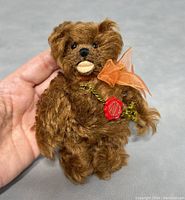 Front view of the small brown teddy bear held in hand to show size and detail, featuring orange bow and red wax seal tag.