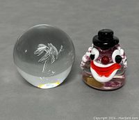 Photo showing clear Danish Holmegaard crystal paperweight next to colorful Italian Murano clown paperweight on gray surface.