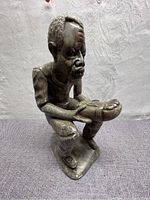 Full view of the soapstone carving of the man sitting, holding and playing a thumb instrument. Shows detailed facial features, textured hair, and clothing folds.