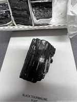 Close-up image of a single flat, rough black tourmaline stone showing the striated layered texture and glossy black surface.