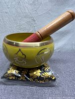 Yellow metal singing bowl with engraved symbols on black and gold floral cushion and wooden mallet inside