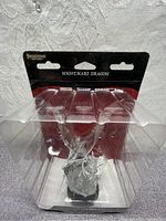 Front view of the Nightmare Dragon miniature in clear plastic blister packaging with black and red cardboard backing showing the name 'Nightmare Dragon' and 'Pathfinder Deep Cuts'. The grey unpainted model is visible.