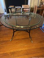 Full view of the round glass top table showing the clear glass surface and metal frame with brass trim and claw feet.