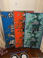 Three fabric art panels leaning against a wall, one blue, one red-orange, one blue-green with medieval motifs.