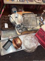 Overview photo showing muffin pans, aluminum and copper Bundt pans, wire cooling racks, large pans, and some bakeware stacked on a table.