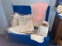 Blue tote filled with various blankets and packaged linens including crocheted and cashmere blankets and new sheet sets.