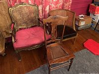 Set of three vintage wooden chairs in room with wood floor and red patterned curtains. Includes ornate armchair with cane back and red cushion, wooden dining chair with carved back and damaged cane seat in front, and barrel chair with cane back and dark upholstery at side.