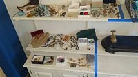 Two white shelves displaying assorted necklaces, brooches, rings, earrings and a black tray
