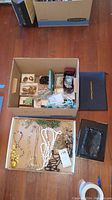 Two open cardboard boxes displaying assorted costume jewelry on hardwood floor