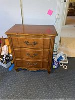 Front view of the Fancher wooden nightstand showing three drawers with decorative metal handles and curved edges, scratches visible on wood surface.