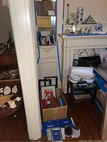 Various picture frames stacked and framed pictures on a narrow white built-in shelf near a fireplace mantel, including multiple frames of different sizes and materials, some in a box and some stacked
