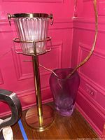 Photo showing both the standing ice bucket on brass stand and the thick purple glass vase with branches inside, placed against a pink paneled wall.