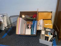 Wide view showing a cardboard box filled with spiral notebooks, legal pads, a decorative note cube, pens, scissors, manila envelopes, and black wire mesh organizers adjacent.