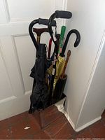 Photo of the lot showing several walking canes and black umbrellas in a corner, displaying various types including with foam and wooden handles.