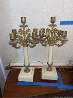 Pair of vintage brass and marble candelabras standing on a wooden surface against a neutral wall. Both have five candle holders and square marble bases.