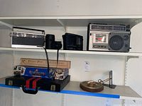 Photo showing shelf with various vintage electronics and boxed board games including Sony transistor radio, Info-Mate world band radio, GE cassette recorder, tavern pipe and games.