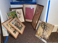 Seven framed wall art pieces leaning against a wall on carpeted floor, various styles and sizes