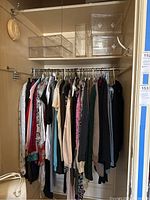 Closet rod with approximately 25 assorted women's clothes on hangers including sweaters, blouses, dresses, and pants; acrylic storage containers on shelf above.