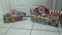 Full view of both Coca Cola themed vintage wall clocks showing soda shop and drive-in scenes on tiled floor.