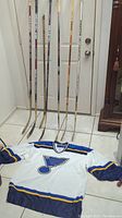 Photo showing seven vintage wooden hockey sticks leaning against a wall with a closed door and a white St Louis Blues jersey laid out on the floor.