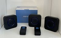 Three black wireless doorbell receivers with central musical note icon illuminated by blue LED ring, two wireless push buttons, and original box in background.