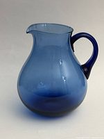 Blue glass pitcher with broad rounded body, narrow neck and large handle, shown against a plain background.