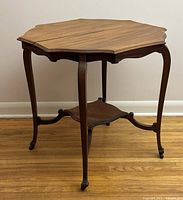 Front view of the solid wood octagonal table on casters standing on wooden floor showing carved edge and cabriole legs with connecting lower shelf.