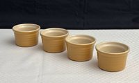 Photo showing four beige/tan La Creuset ramekins in a row on white surface with black background.