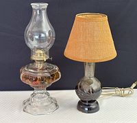 Photo of both oil lamp and ceramic lamp with a brown fabric shade placed side by side on a white surface against a black background. Both lamps are visible with oil lamp on left, showing its clear glass and brass components.