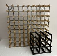 Large wine rack with metal frame and wood pegs holding 64 bottles next to small black plastic rack for 9 bottles on a white surface, front view.