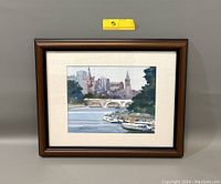 Framed watercolor painting of a cityscape with bridge and riverboats hung on a wall with a yellow lot number tag above it.