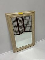 Rectangular wooden framed wall mirror with carved decorative details on the frame, light wood color, reflecting room divider in background.
