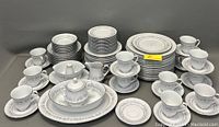 Full set showing cups on saucers, stacked plates (dinner, bread, salad), dessert and soup bowls, sugar bowl with lid, creamers, vegetable bowl, and platter, all with a light blue and grey floral pattern and silver trim.